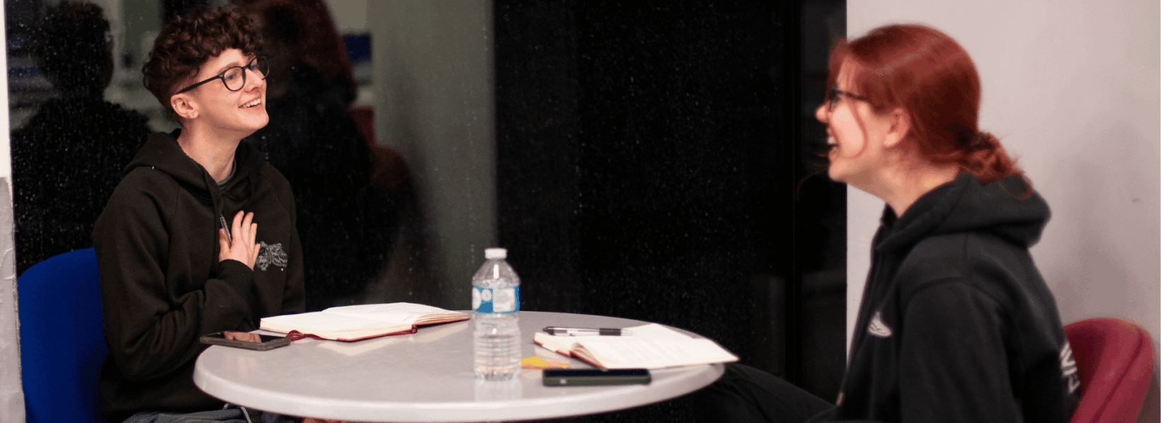 Two young people smiling and talking together during a creative workshop, seated at a table with notebooks.